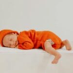 Śpiący noworodek w pomarańczowym śpioszku na małym białym łóżeczku