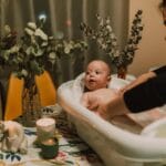 Mama kąpie niemowlę w małej wanience wśród ciepłego światła i roślin