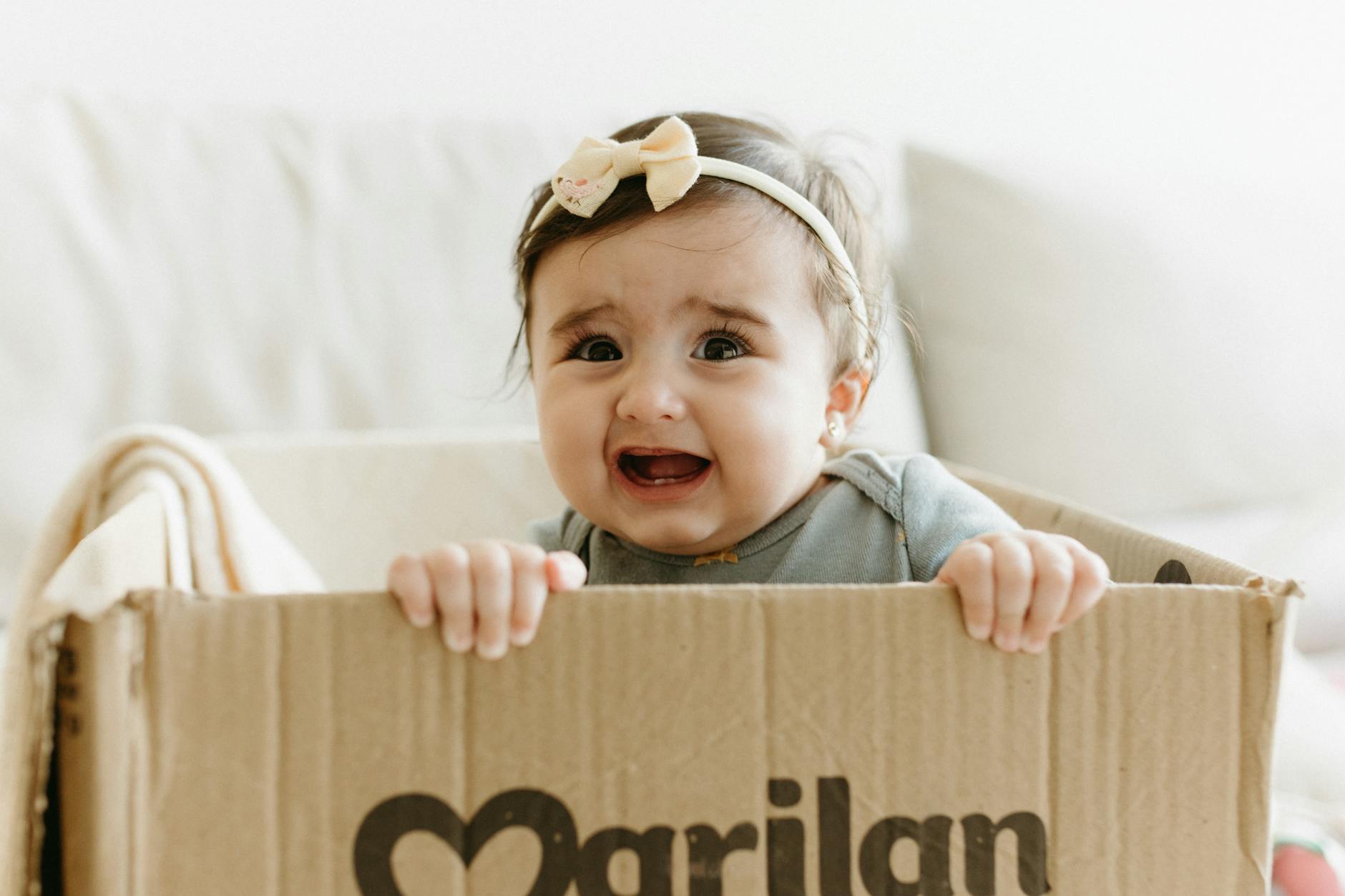 Niemowlęca dziewczynka z kokardką bawi się, siedząc w kartonowym pudełku