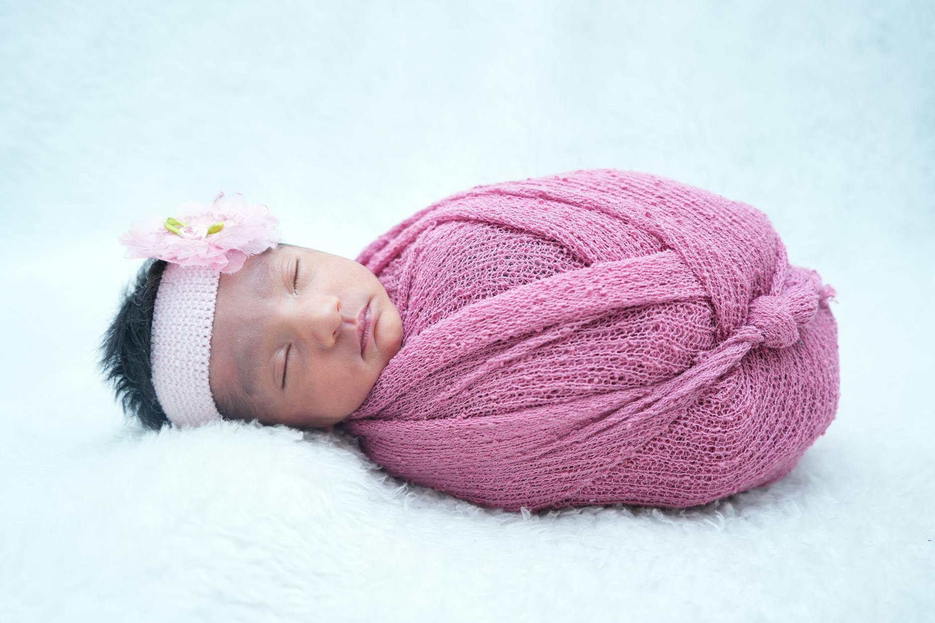 Noworodek owinięty różowym kocykiem, śpiący z kwiatową opaską